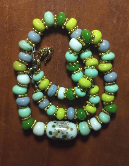 A handmade lampwork glass bead necklace in shades of opaque green and grey, set against a dark wood background.
