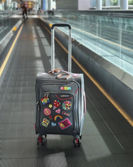 A wheeled suitcase with various colorful stickers and patches, displaying the word "ALOHA" and images like flowers and rainbows, is placed on an airport moving walkway. The modern, spacious terminal is visible in the background.
