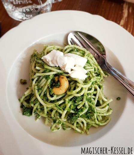 Draufblick auf einen weißen Porzellanteller mit Spaghetti, die von frischem, frühlingsgrünen Pesto ummantelt sind. Obendrauf liegen einige Stücke Mozzarella und eine geröstete Cashew. Oben rechts im Teller liegen ein Löffel und eine Gabel.