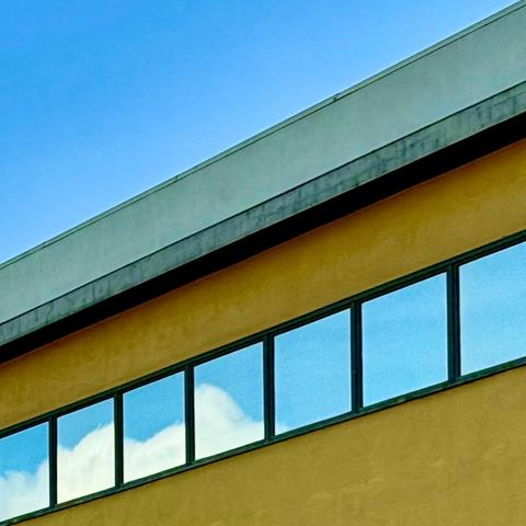Diagonal shot of the top of a yellow commercial building with a row of windows with black panes. The windows are all reflecting a blue sky, some with white puffy clouds. Above the building is a clear blue sky.