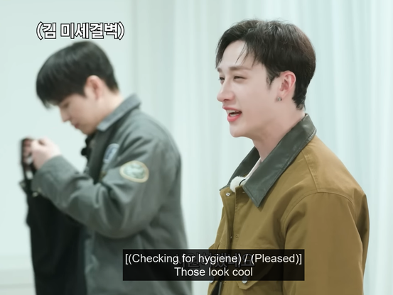Bang Chan in the foreground saying, "Those look cool." In the background, Seungmin holding up the black tank top and investigating it. Caption: [(Checking for hygiene) / (Pleased)]