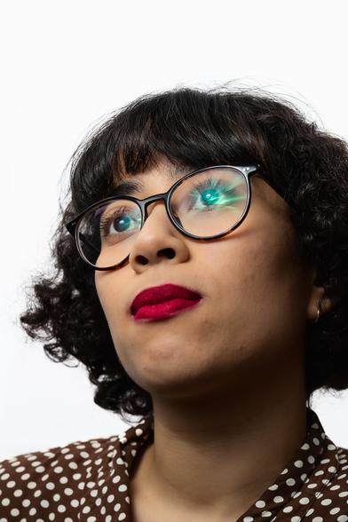 Head-and-shoulders shot of a person with short, curly hair, wearing round, dark-framed glasses. They are gazing upward, and the lenses of the glasses reflect a soft light. Their lips are painted a vivid red. The background is plain white, with the bright lighting focusing on the person’s face.