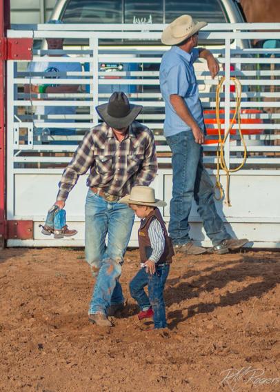 Photograph of a father and young son walking thru the dirt of a rodeo arena, the father carrying the son's cowboy boots in one hand while holding his hand.