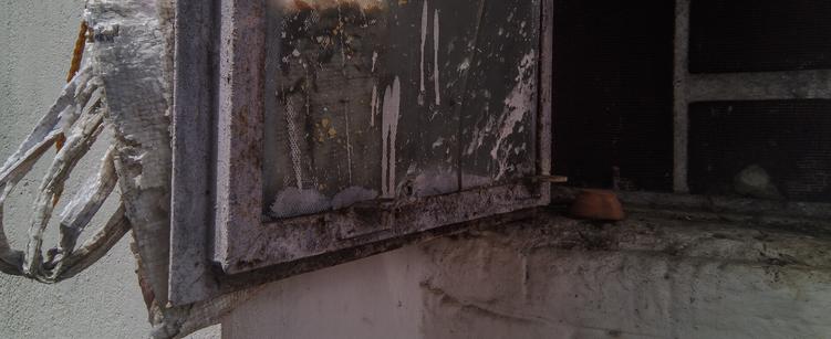 A wide cropped photo of a window and a windowsill. The window is made out of metal painted white, and the paint is chipping and the metal is rusting through the white paint. The glass in the window has white splashes of paint on it and is frosted. Behind the window, there is a white carry bag made out of white plastic, which is visible through the window pane as well. On the window sill, there is an earthen lamp in earthen unpainted colour.