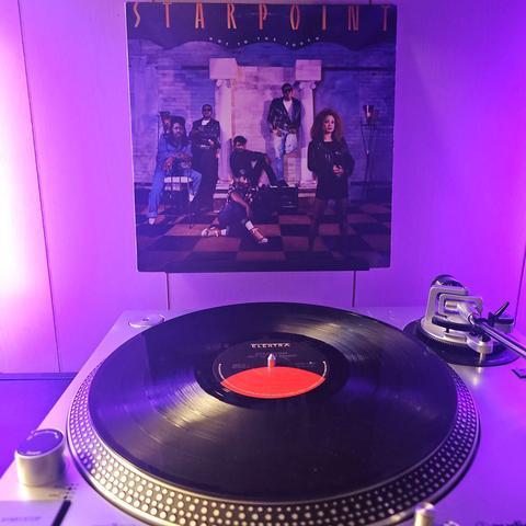 A vinyl record sits on a turntable. Behind the turntable, a vinyl album outer sleeve is displayed. The front cover shows the members of Starpoint (5 men, and 1 woman) in front of a brick wall and pillars posing for the camera.