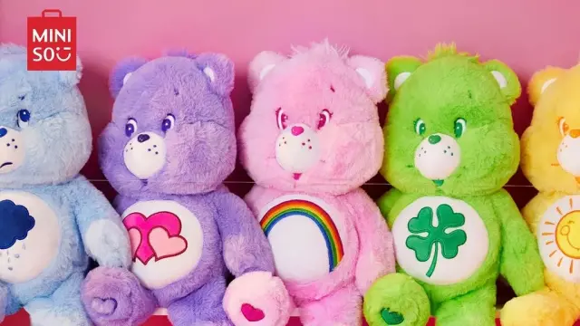 Photo de cinq peluches Bisounours de différentes couleurs (bleu, violet, rose, vert, jaune) alignées. Le logo des boutiques Miniso apparaît dans le coin.