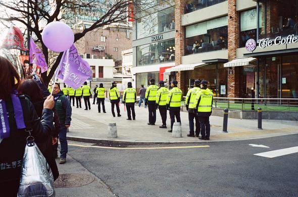 형광 유니폼을 입은 경찰들이 인도에 길게 늘어서있다. 왼편에는 보라색으로 꾸민 민주노총 조합원들이 행진하고 있다. 오른편 카페 건물에서 시민들이 민주노총 행진을 내려다보고 있다.