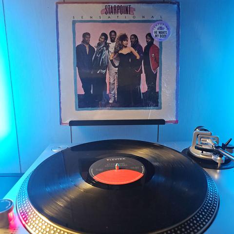 A vinyl record sits on a turntable. Behind the turntable, a vinyl album outer sleeve is displayed. The front cover shows the members (5 men and 1 woman) standing together looking at the camera.