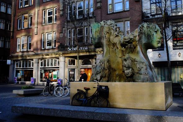 Man smoking pipe, leaning against statue, in front of bookshop, bicycles all around, patches of highlights from reflection of sun in windows at opposing side of the street.