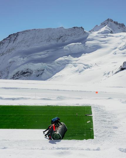 The image shows a snowy mountain landscape with people rolling out a large section of artificial green turf over the snow. The backdrop includes snow-covered peaks under a clear blue sky.