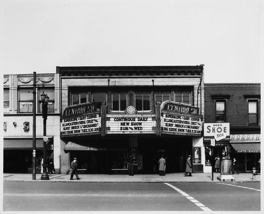 A black-and-white photograph of a movie theater shot from across Mass Ave (viewer facing northeast).  The side panels of the theater marquee read 'Central Sq' over the featured films: 'Superscope - Gary Cooper - B. Lancaster in "Vera Cruz" | also Elroy Hirsh in "Unchained" | Big Kiddie Show Tues 9:30 AM'.  The center panel reads 'CONTINOUS[sic] DAILY NEW SHOW SUN and WED'.

On the left side of the frame one can see part of Gorin's department store, above which is the office of V Multer, dentist. On the right side of the frame is Bob's Shoe Box, to the right of which is a hat and bag store called Dorothy's.  Several pedestrians are visible on the sidewalk.