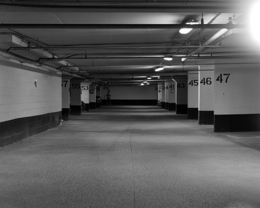 The view from my parking spot: a low concrete ceiling, broken up but the regular patterns of the sprinkler piping. Fluorescent lighting recedes to the end of the row with the space dividers numbered in stencilled black pain.