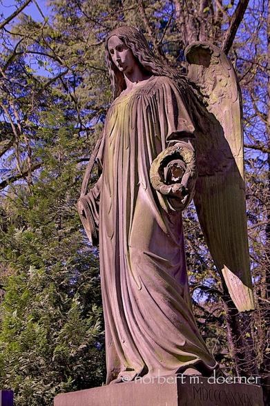 Steinerne Engelsfigur mit zwei Schwingen, aber schon abgefallenen Händen, und leeren Augenhöhlen auf einem Podest vor einem Baum und blauem Himmel.
