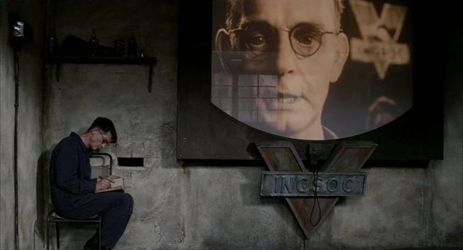 A man sitting sideways on a chair, tucked tightly in a corner. The brick is removed from the wall and he is writing in a diary. A giant screen looms over him, with a man’s close-up face. There is a sign reading “INGSOC”.