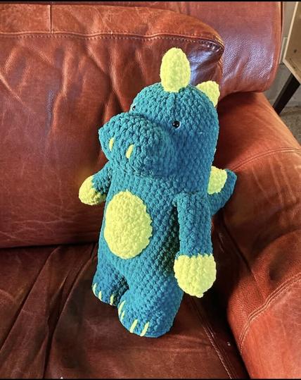 A blue and bright lime green stuffed dinosaur propped on a red, leather couch. The dinosaur stands upright and has lime green spikes on its back with green hands, belly plate and embroidered details for nostrils and toes. It has black safety eyes.