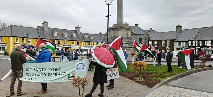 About thirty people on the traffic island at the Octagon in Westport, Co. Mayo, Ireland, holding Palestinian flags and banners, one person with a watermelon umbrella, buildings in the background