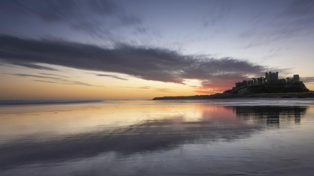 A blue hour photo of a castle standing next to a beach. The sand on the beach is wet from the occasional wave that manages to reach quite a long way in land as the sand is so flat. The sun is rising behind the castle which is silhouetted against the colourful  sky. The cloud which is being lit up by the sun is bright red and it resembles a fire with smoke.

A woman walks her dog on the beach