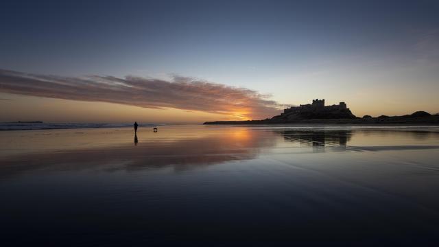 A blue hour photo of a castle standing next to a beach. The sand on the beach is wet from the occasional wave that manages to reach quite a long way in land as the sand is so flat. The sun is rising behind the castle which is silhouetted against the colourful  sky. The cloud which is being lit up by the sun is bright orange.

A woman walks her dog on the beach