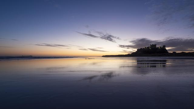 A blue hour photo of a castle standing next to a beach. The sand on the beach is wet from the occasional wave that manages to reach quite a long way in land as the sand is so flat. The sun is rising behind the castle which is silhouetted against the colourful  sky.