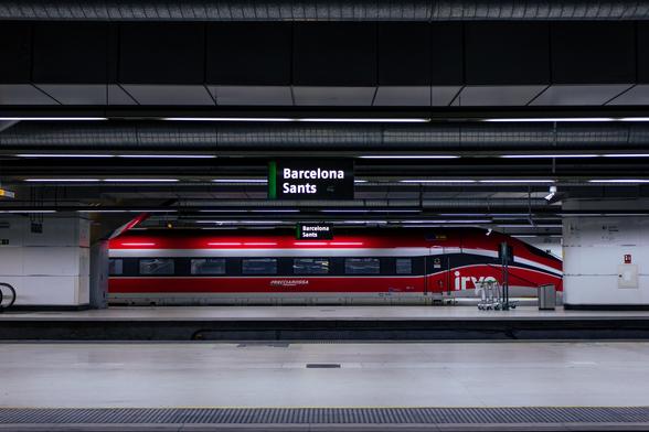 A striking red ETR 1000 "Frecciarossa" high speed train, stationed at Barcelona Sants train station. The picture, captured from several platforms away, shows the side of the train, which highlights the aerodynamic design the train has.