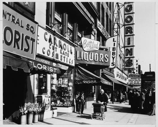Black and white photograph of an urban sidewalk, featuring storefronts labeled "Central Sq Florist", "Carroll Cut Rate Cosmetics", "Libby's Bottled Liquors", "Gorin's", "Central Sq" (Theatre) and others.  The theatre marquee advertises "Adventurous piracy - Cinemascope - Robert Newton as 'Long John Silver' - and Glenn Ford in 'Americano'".  Two preadolescent children loiter in front of the liquor store; a woman pushes a pram; pedestrians crowd the MBTA station entrance, which is marked "Trains for Harvard Square".