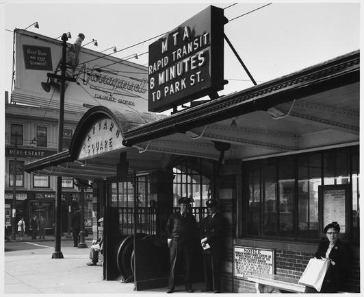 Black and white photograph of the Harvard Square train station entrance.  The sign overhead reads "MTA RAPID TRANSIT - 8 MINUTES TO PARK ST."   Two uniformed white men under the awning smile for the camera.  A large billboard advertisement for Narragansett lager beer dominates the background; other pedestrians are visible on the sidewalk.