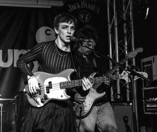 Black and white photo of male bass player at a gig. He is wearing a black top and eyeliner