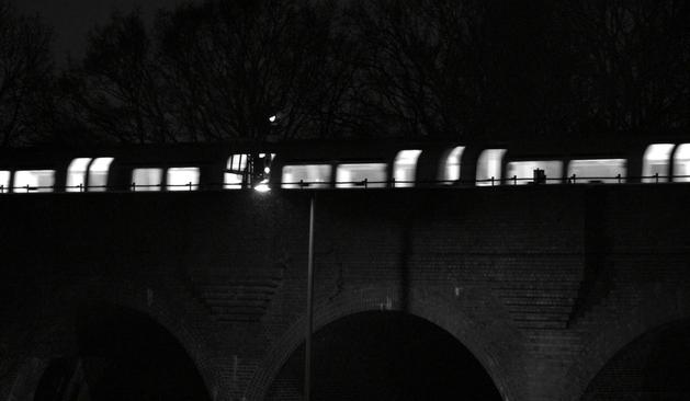 A subway glides across the brick viaduct at night, its motion blurred in the moonlight. Silhouetted trees form a dark backdrop, while a streetlight softly illuminates part of this over-ground route.

Eine U-Bahn gleitet ueber das naechtliche Backsteinviadukt, ihre Bewegung verschwimmt im Mondlicht. Silhouettierte Baeume bilden einen dunklen Hintergrund, waehrend ein Strassenlicht einen Teil dieser oberirdischen Strecke sanft beleuchtet.