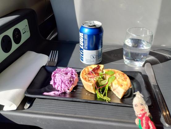 A seat back table on a train. Brie and red onion quiche served with artistic salad and a red cabbage coleslaw. A glass of sparkling water. A finger puppet.