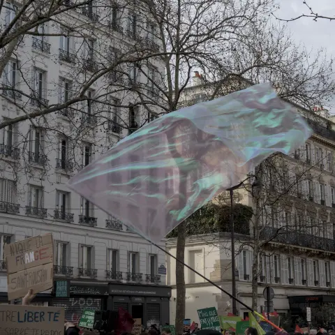 Photograph of a large, transparent flag of pink-ish color with green-ish reflections being waved over protestors' heads at a march in Paris.