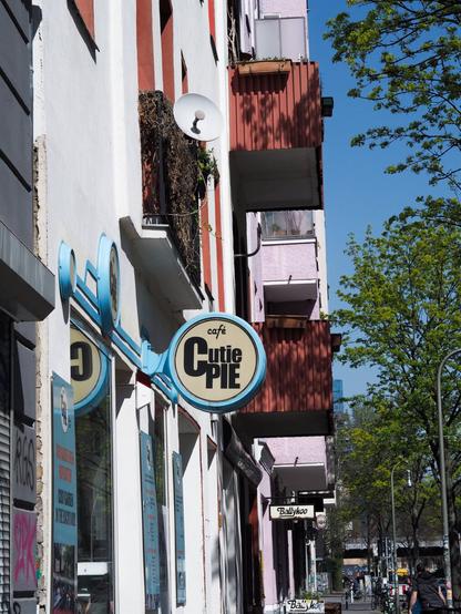 Picture of a street in Berlin. In the focus of the photo there is a round sign of a coffee shop attached to a building. The sign has cafe's name which is Cutie Pie. The weather is sunny, sky is blue and trees next to the sidewalk are green with new leaves.
