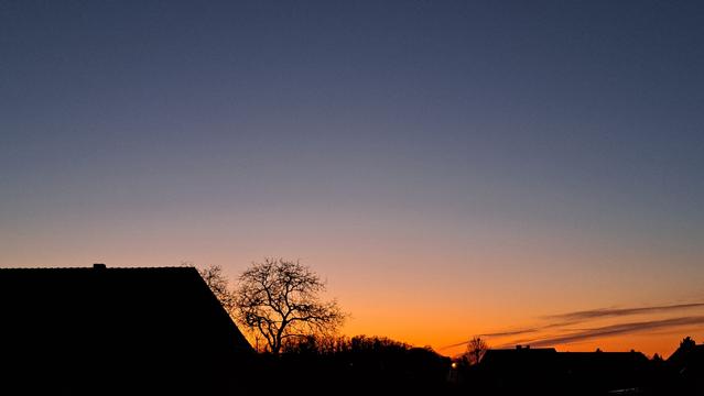 Fensterblick: Dunkelblauer Abendhimmel mit einem leuchtenden Streifen Orangerot überm Horizont sowie ein paar wenigen, streifigen Schleierwolken. Silhouetten von Hausdächern und Bäumen am unteren Bildrand