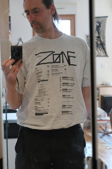 A man with a digital camera shooting himself in the mirror, he wears a gray tee shirt with various lettering, lyrics, and custom-made icons.