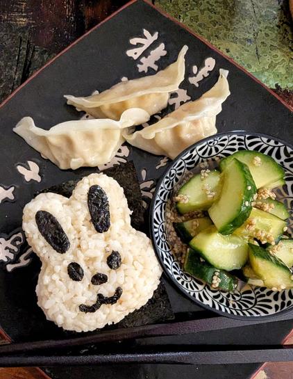 Dark wooden table, black asian dinner plate. On the plate: a bunny onigiri, the face is made with cut out nori bits. alongside is a small bowl, filled with smashed cucumber salad with a brownish dressing full of sesame seeds. You can also see 3 shrimp gyoza and a pair of black chop sticks on the plate.