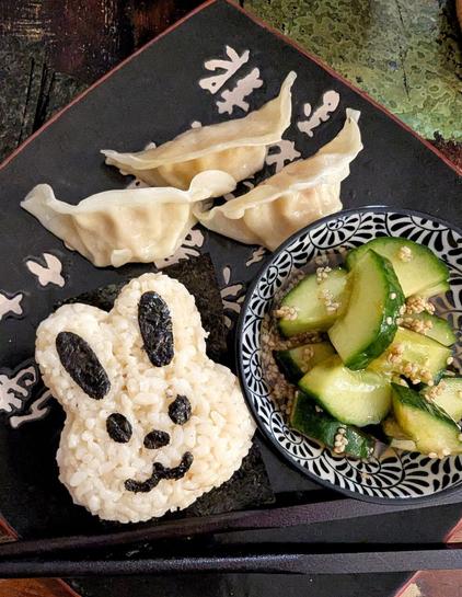 Dark wooden table, black asian dinner plate. On the plate: a bunny onigiri, the face is made with cut out nori bits. alongside is a small bowl, filled with smashed cucumber salad with a brownish dressing full of sesame seeds. You can also see 3 shrimp gyoza and a pair of black chop sticks on the plate.
