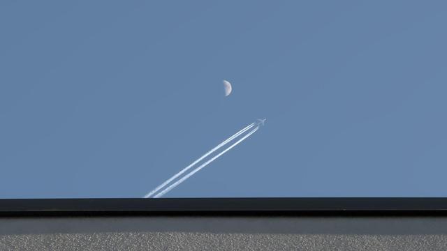 Abstract looking photo: white and black roof in the bottom, light blue sky above it. In the sky, a plane with a trail is passing the half moon.