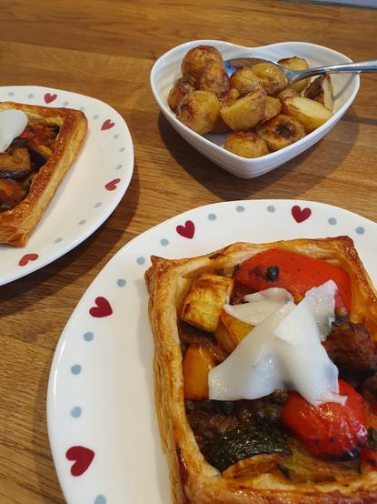 The image shows two rectangular puff pastry dishes topped with colorful roasted vegetables and slices of cheese, served on plates with heart designs. A small white heart-shaped bowl contains golden roasted potatoes with a spoon. The background features a wooden table.