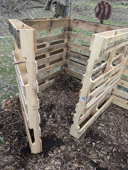 Four shipping pallets have been tie-strapped together, in the corner of a backyard (garden) to form a composting bin. One of the pallets is partially open to demonstrate the gate that allows access.