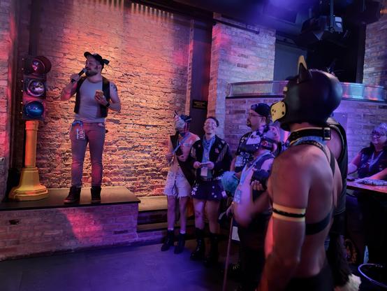 A vibrant nightlife scene unfolds in a dimly lit bar with exposed brick walls bathed in dramatic red and purple lighting. On a small stage, a performer in a white tank top, vest, and red pants holds a microphone while checking their phone, seemingly hosting or performing at an event. In the foreground, an attentive audience watches, including a person wearing a distinctive black leather puppy hood with yellow accents and harness straps across their bare torso. The crowd appears diverse and enthusiastic, with several attendees wearing alternative fashion including various leather accessories and harnesses. The intimate venue creates a warm, underground club atmosphere with its brick walls and theatrical lighting, suggesting this might be an opening night or special event for a community gathering. The combination of performance space, specialized gear, and engaged audience creates a sense of belonging and celebration within this alternative nightlife scene. #LeatherCommunity #NightlifeScene #AlternativeFashion #PuppyPlay #ClubNight