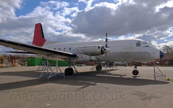 Side view of a white, twin propellor-engined airliner, facing to the right, suspended on white metal supports under the wings and fuselage.
The white fuselage has paint stripped off the belly with the remnants of red paint on the bottom of the nose cone, with the red tail almost hidden behind the wing.
Grey concrete fills the lower half of the frame, with a strong shadow being cast on the ground.
Green storage containers and trees are visible under the planes body, with blue sky and scattered fluffy white cloud above.