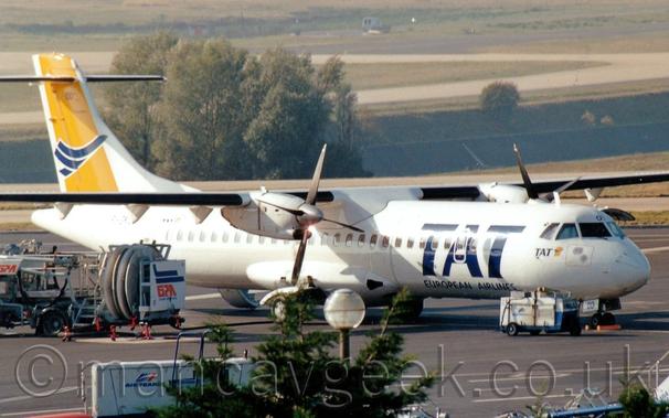 Side view of a white, high-winged, twin propellor-engined airliner parked facing to the right.
There are large, billboard-style "TAT" titles on the forward fuselage, above smaller "European Airlines" titles.
There are smaller, blue "TAT" titles under the cockpit, next to a small, yellow square with a stylised blue flying bird.
This same logo appears in a larger form on the tail, 
The registration "F-GKOD" is on the upper rear fuselage in blue, almost obscured by bright sunlight reflecting off the fuselage.
A tree is in the middle of the frame at the bottom, with a white truck behind it.
A refueling truck is on the left of the frame, in front of this plane.
Trees and large grassed areas with taxiways and a runway fill the rest of the background.