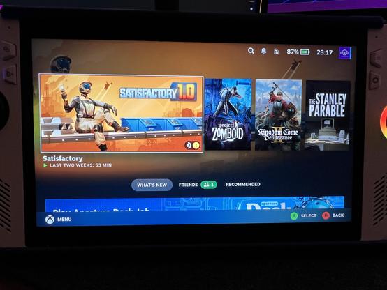 The image shows a gaming console screen displaying a game library. The screen is divided into sections, with the left side featuring the game "Satisfactory" prominently, showing a character in a space suit sitting on a platform with the text "Satisfactory 1.0" and "Last Two Weeks: 53 Min" below it. The right side displays three other game covers: "Project Zomboid," "Kingdom Come: Deliverance," and "The Stanley Parable." The top of the screen shows the time as 23:17, the battery level at 87%, and a notification icon. The bottom of the screen has navigation options labeled "What's New," "Friends," and "Recommended," with a "Menu" button on the left and "Select" and "Back" buttons on the right. The overall color scheme is dark with blue and orange accents.

Provided by @altbot, generated privately and locally using Ovis2-8B

🌱 Energy used: 0.207 Wh