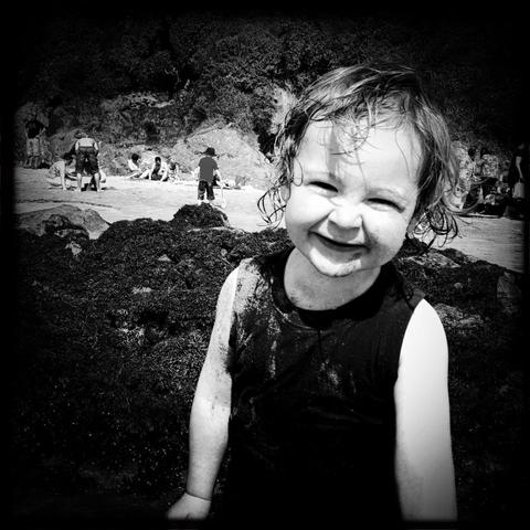 A black and white photo of a smiling child at the beach, covered in sand. The child has wet, curly hair and is wearing a sleeveless shirt. In the background, people are relaxing on the sand and rocky areas.