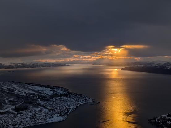 The sun still hiding behind a cloud lights up an orange streak over the fjord towards the camera while another part of the fjord is silver from the sunlight reflected of the sky/clouds.