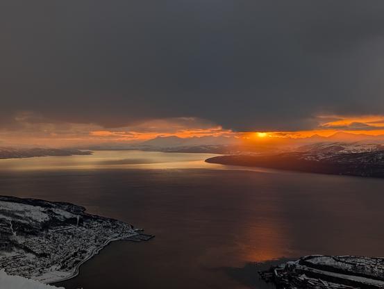 A sun peaks from under a grey cloud cover. While lighting up the fjord to the left. Dark orange with silver.