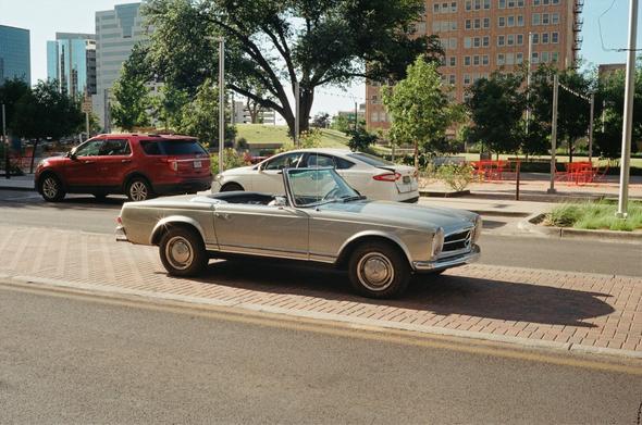 Older convertible Mercedes SL parked in the middle of a street in front of a park.