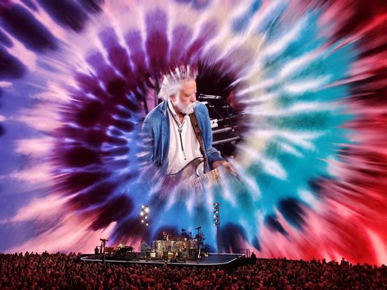 Bob Weir, encircled by a tie-dye design, projected on a giant screen above the band Dead and Company performing in front of an audience.