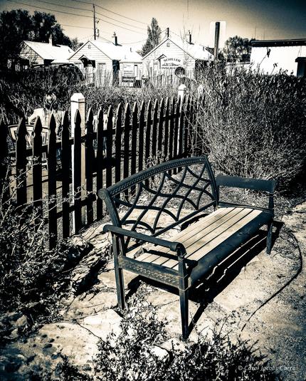A split tone green and yellow monochrome photo of a garden bench on a stone patio. This is a backyard with a picket fence net to another yard.  You can see the houses converted to office across the street,