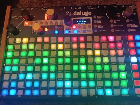 A rectangular music making synth box with a rainbow colored grid of pads, a few knobs, glowing buttons and a small screen