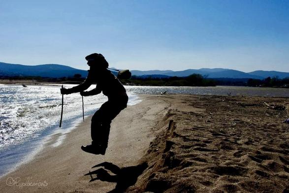 Un estuaire de fleuve, on voit une plage de sable doré au premier plan, la mer sur la gauche, le fleuve qui s’y jette sur la droite, et des collines dans le fond qui se découpent devant un beau ciel bleu. Le sujet de la photo est un homme à contre-jour en train de sauter vers la mer, les deux pieds en avant et les mains tenant deux batons, comme s’il sautait une dune de neige à ski.